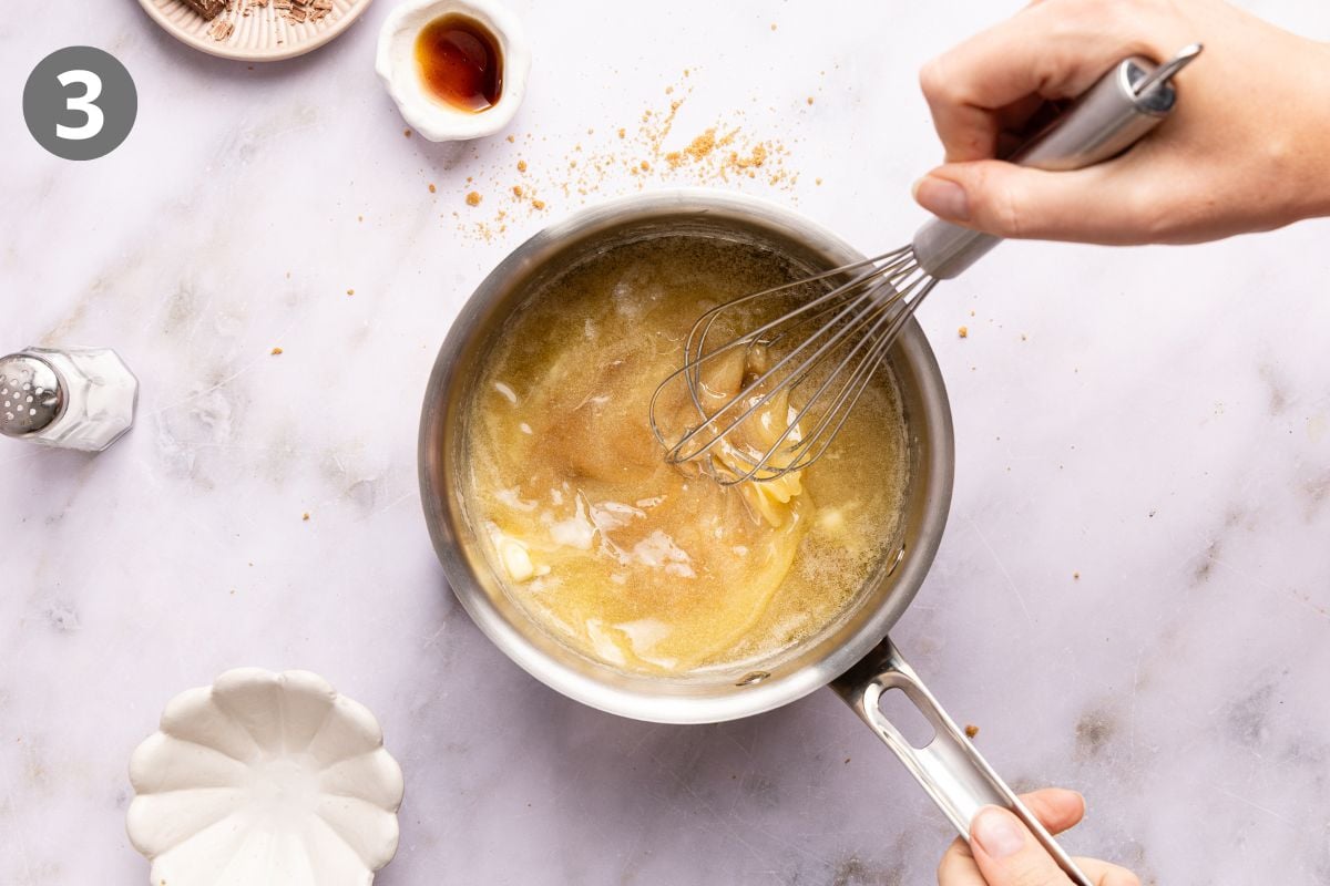 A hand uses a whisk to stir melted butter and sugar in a saucepan.