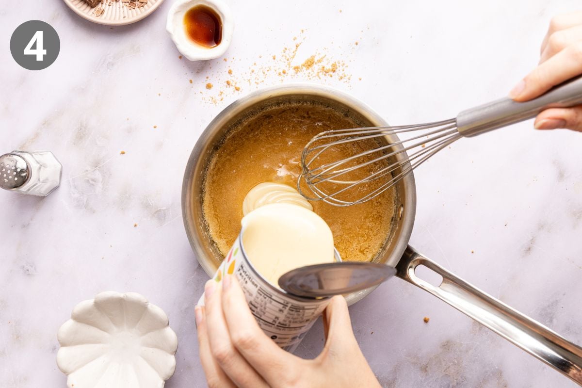 A hand pours a can of sweetened condensed milk into a saucepan containing a melted mixture, while another hand whisks the ingredients together.