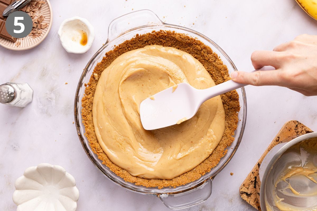 A hand uses a white spatula to spread caramel into a graham cracker pie crust.