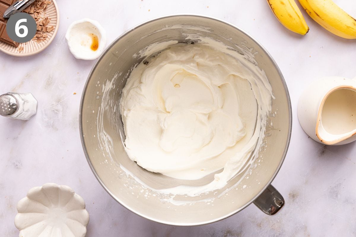 A metal mixing bowl filled with whipped cream, surrounded by bananas, chocolate shavings, and small bowls of ingredients.