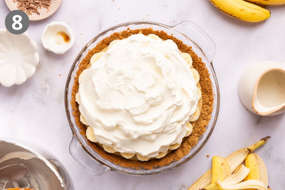 Top-down view of a banoffee pie in a glass dish, topped with a large mound of whipped cream and surrounded by banana slices, a small bowl of vanilla, and banana peels.