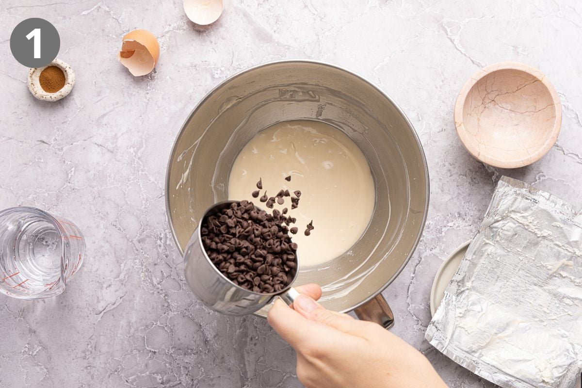 A hand pours a metal measuring cup of chocolate chips into a mixing bowl containing a light-colored batter.