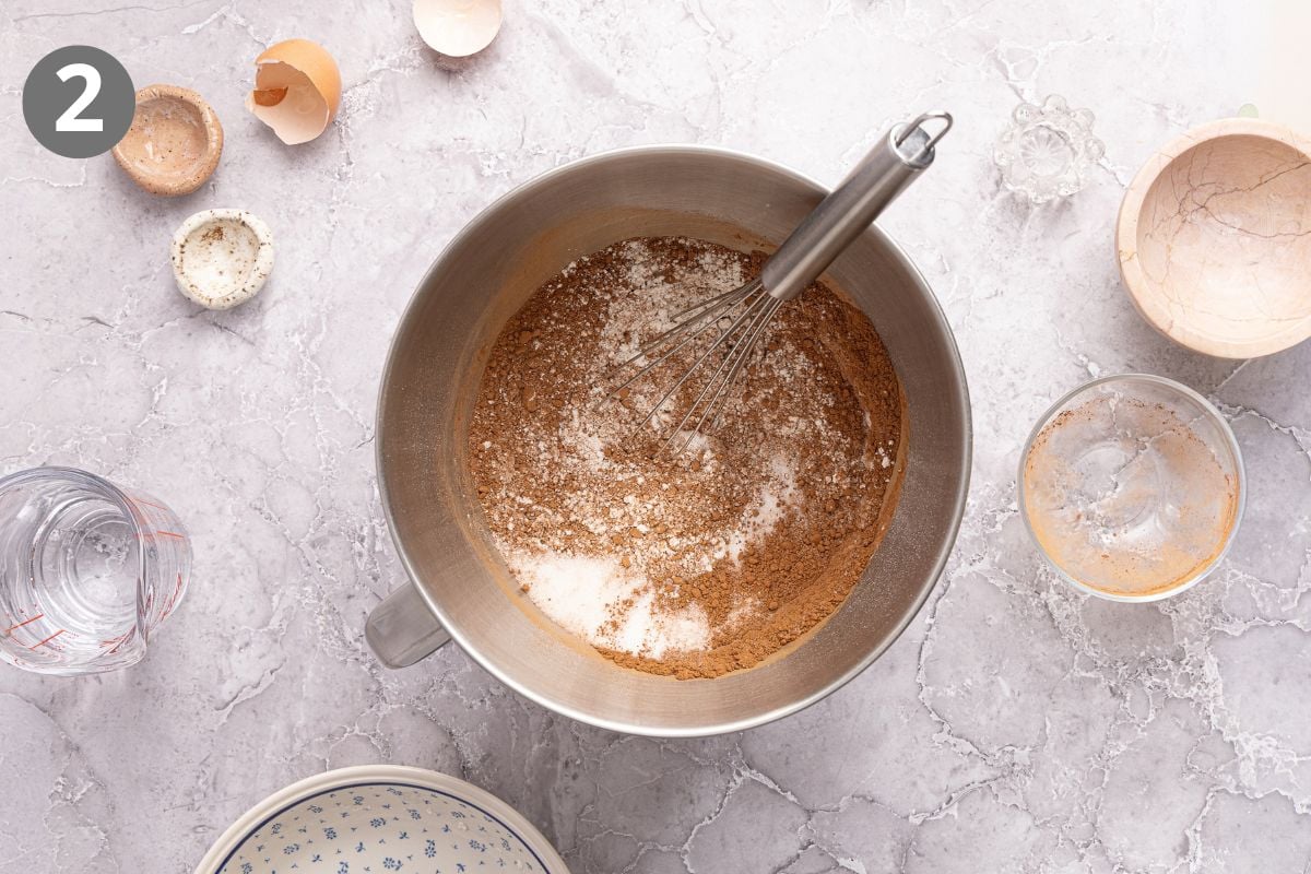 A metal mixing bowl containing dry ingredients including flour and cocoa powder being whisked, surrounded by measuring cups and eggshells.