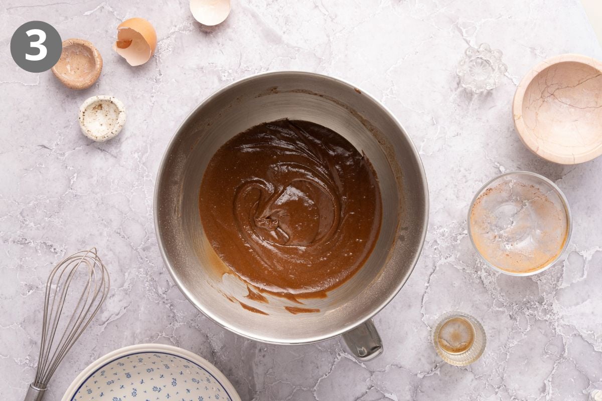 A metal mixing bowl containing smooth chocolate batter, surrounded by a whisk, eggshells, and small bowls of ingredients on a marble countertop.