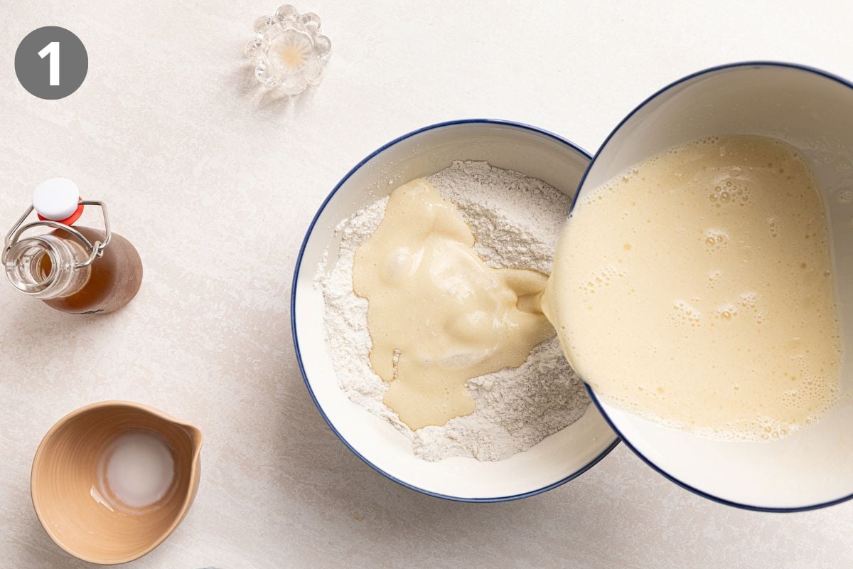 A bowl of dry ingredients being combined with a bowl of wet ingredients, with a small bottle of syrup and a small cup of liquid nearby.