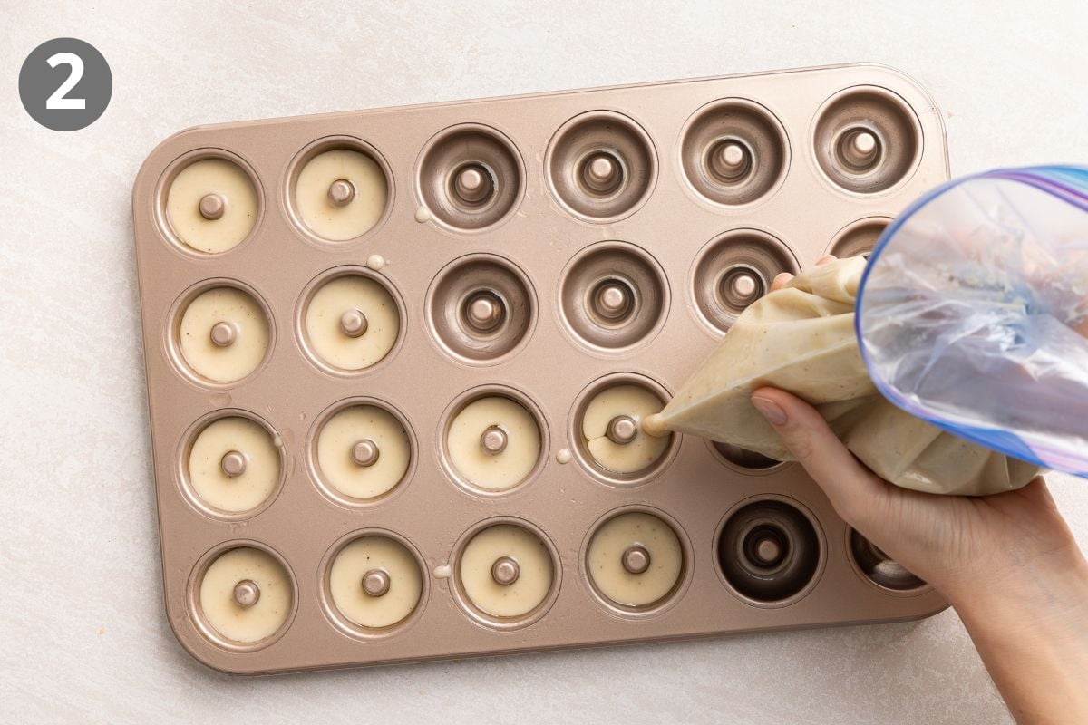 A hand uses a piping bag to fill a metal donut pan with batter.