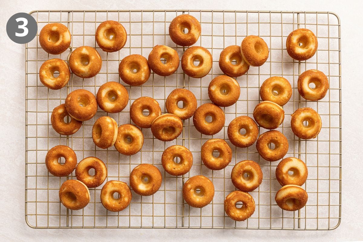 Many small, golden-brown baked doughnuts spread across a wire cooling rack.