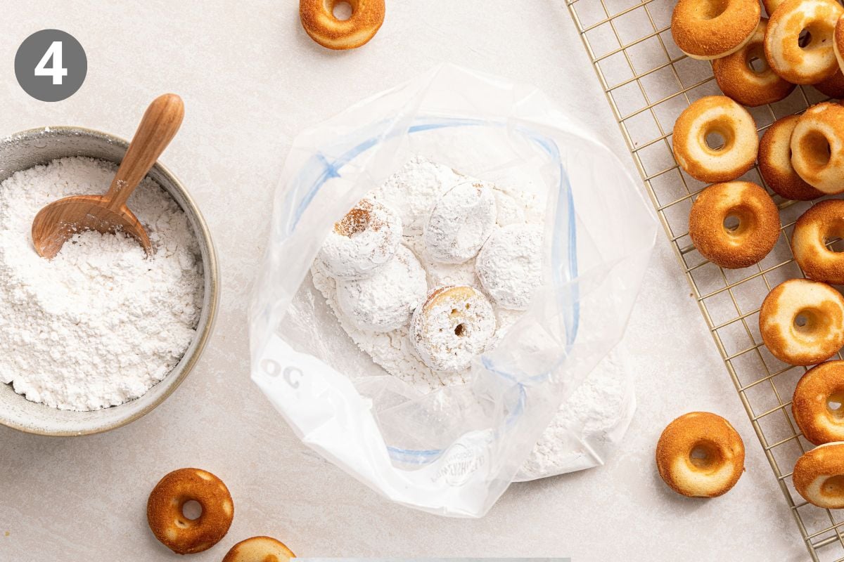 Small donuts being coated in powdered sugar inside a plastic bag, next to a bowl of sugar and a wire cooling rack of plain donuts.