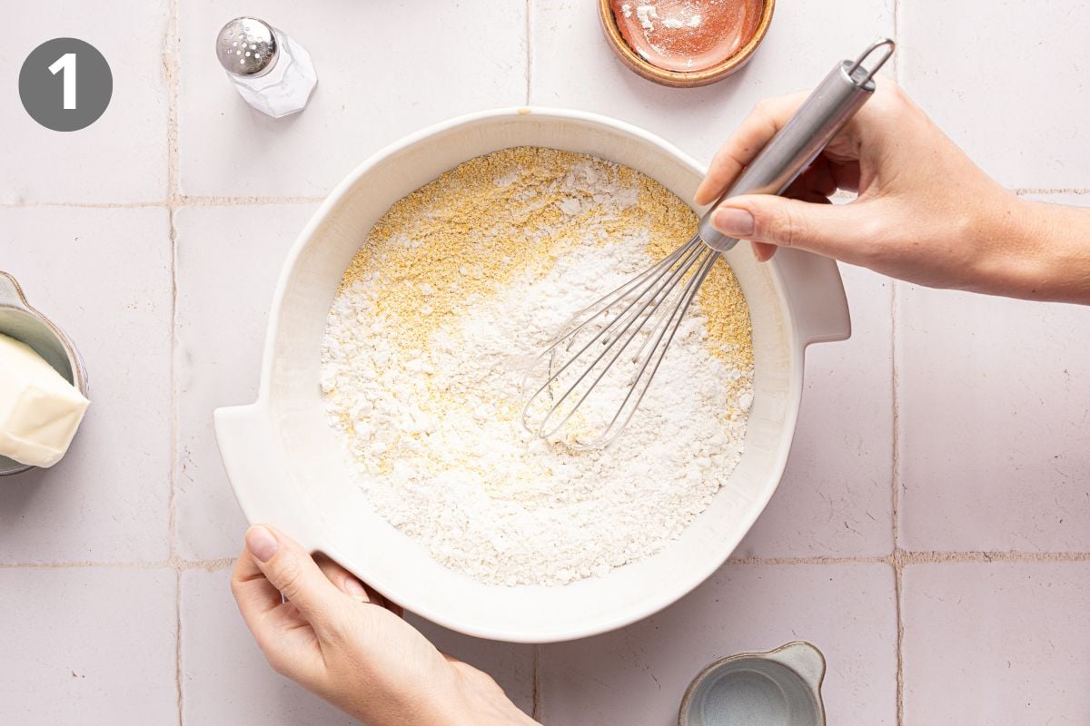 A hand whisks dry ingredients, including flour and cornmeal, together in a white mixing bowl.