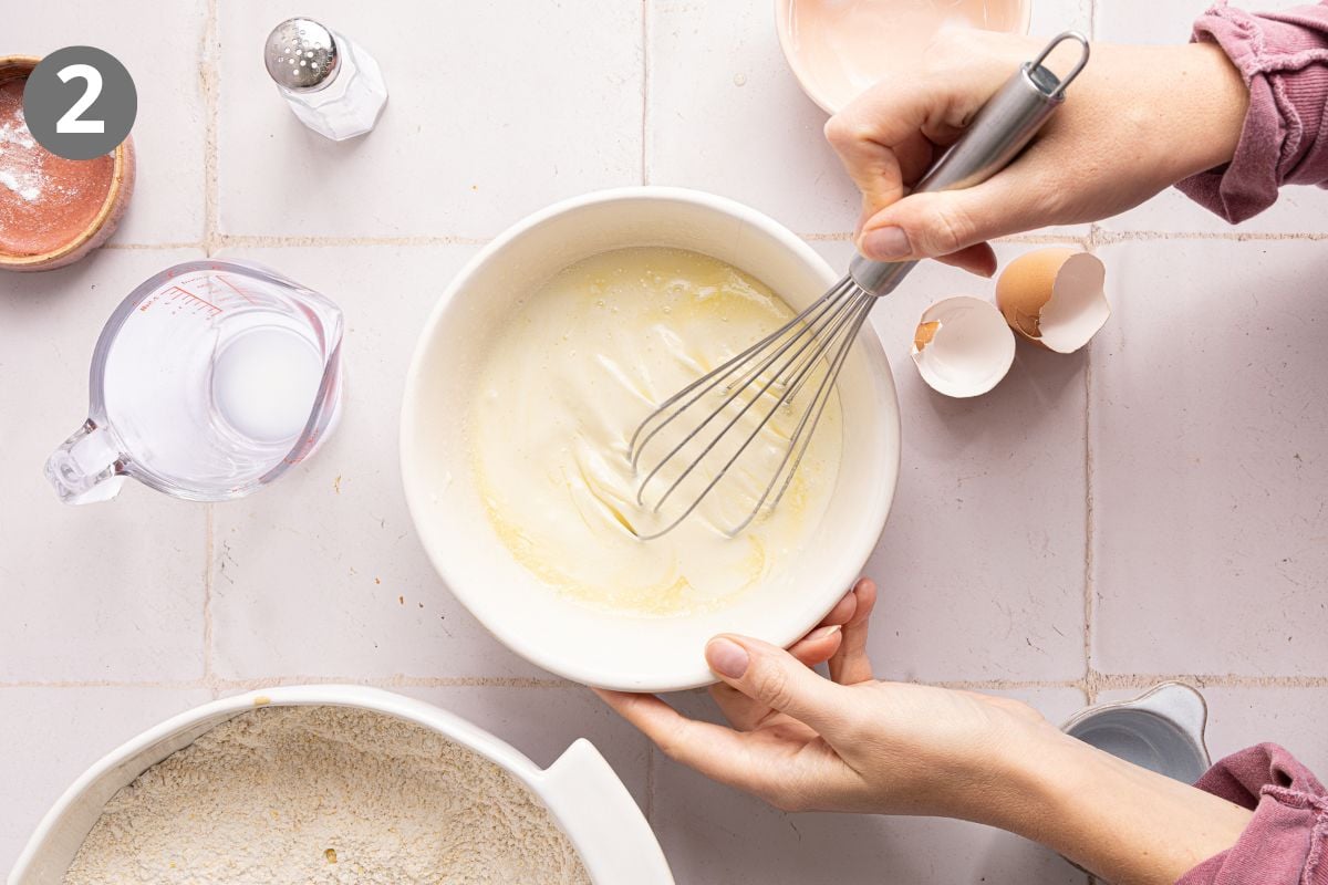 Hands whisking liquid ingredients in a white bowl with a metal whisk, beside a measuring cup of milk and a broken eggshell.