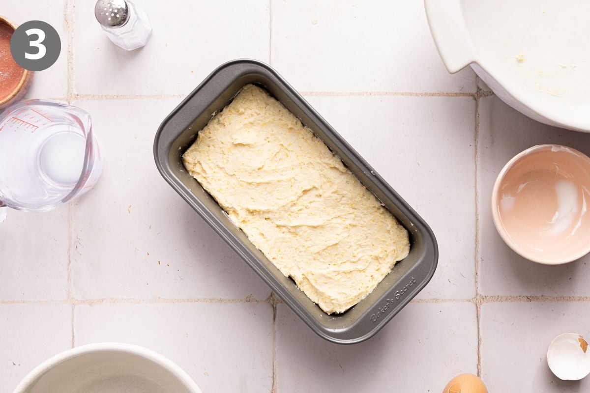 Bread batter in a rectangular metal loaf pan, surrounded by ingredients including a measuring cup of milk, a small bowl, an egg, and a shaker on a tiled surface.