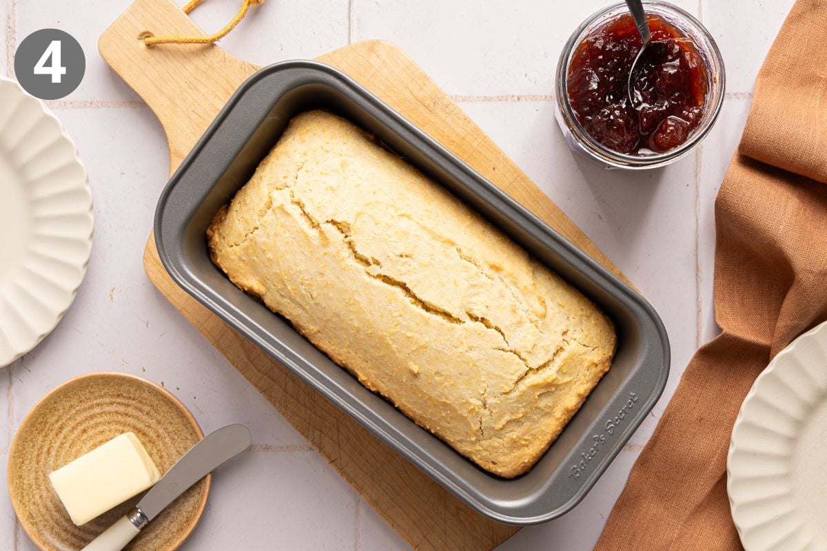 A loaf of cornbread in a metal baking pan, next to a jar of jam, a pat of butter, and two plates.