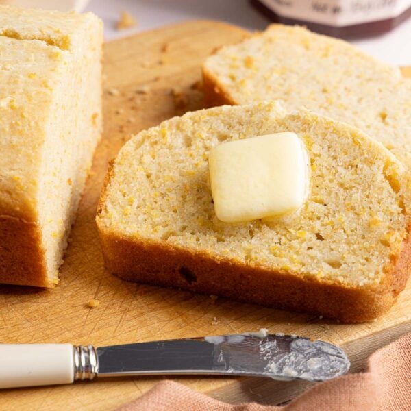 A slice of cornbread topped with a melting pat of butter, sitting on a wooden cutting board next to a butter knife.