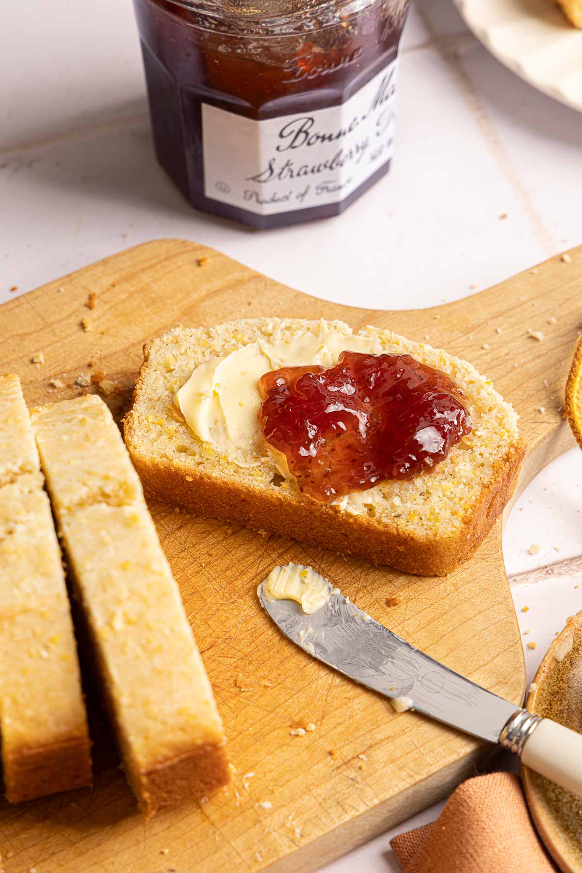A slice of golden bread topped with butter and strawberry jam on a wooden cutting board, with a butter knife and a jar of jam nearby.