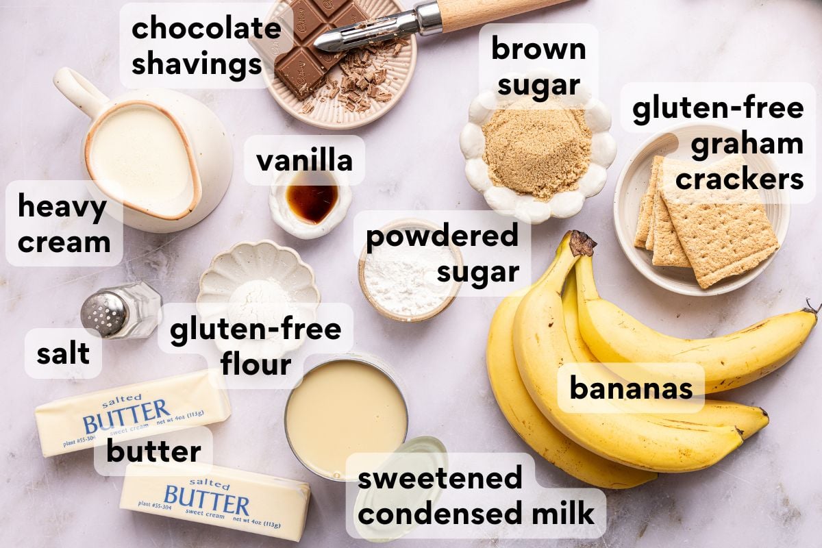 Flat lay of recipe ingredients including heavy cream, chocolate shavings, vanilla, brown sugar, gluten-free crackers, salt, gluten-free flour, powdered sugar, butter, sweetened condensed milk, and bananas, each labeled with its name.