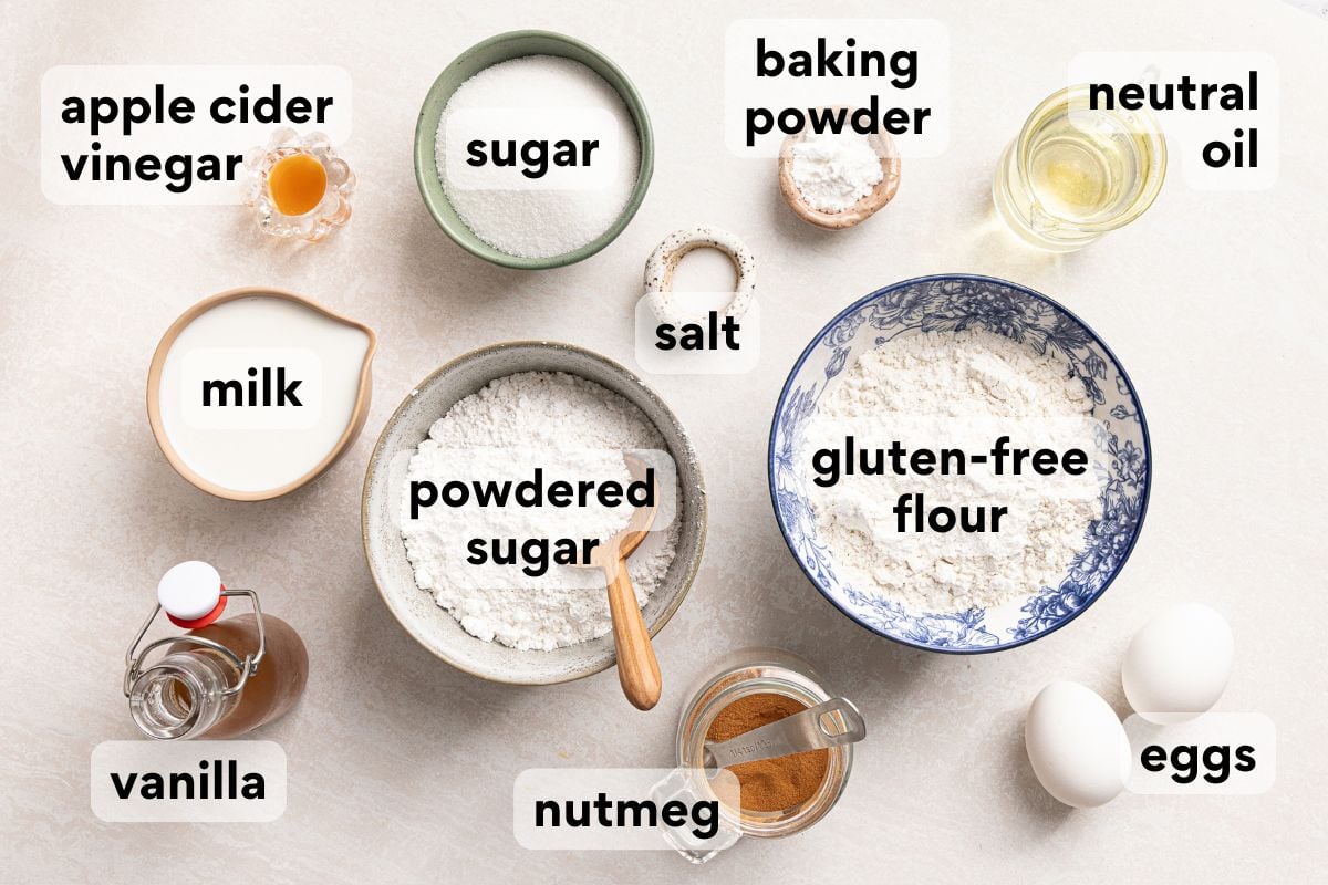 Ingredients for a recipe labeled apple cider vinegar, sugar, baking powder, neutral oil, milk, salt, powdered sugar, gluten-free flour, vanilla, nutmeg, and eggs arranged on a light surface.