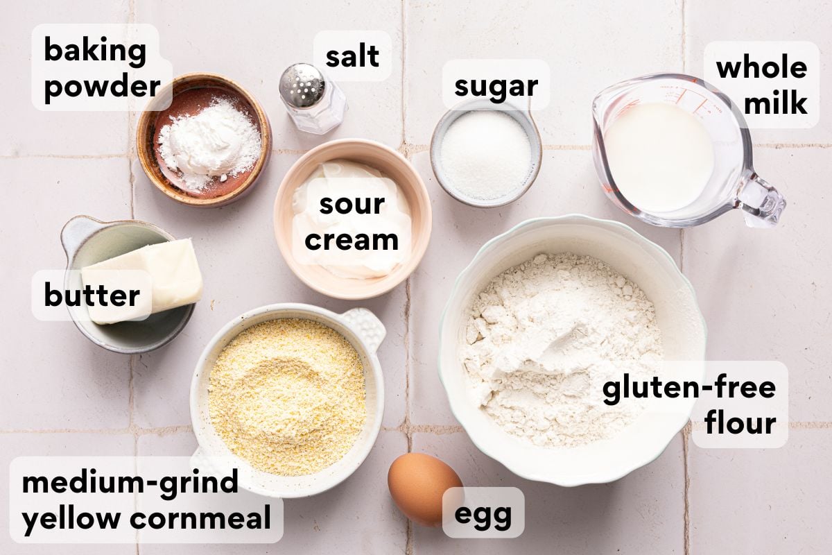Ingredients for a recipe arranged on a surface, labeled: baking powder, salt, sugar, whole milk, butter, sour cream, medium-grind yellow cornmeal, gluten-free flour, and an egg.